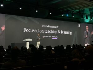 A presenter stands on stage in front of a large screen displaying the text: Who is Blackboard? Focused on teaching & learning. Built with you. Thank you. An audience is seated in the foreground.